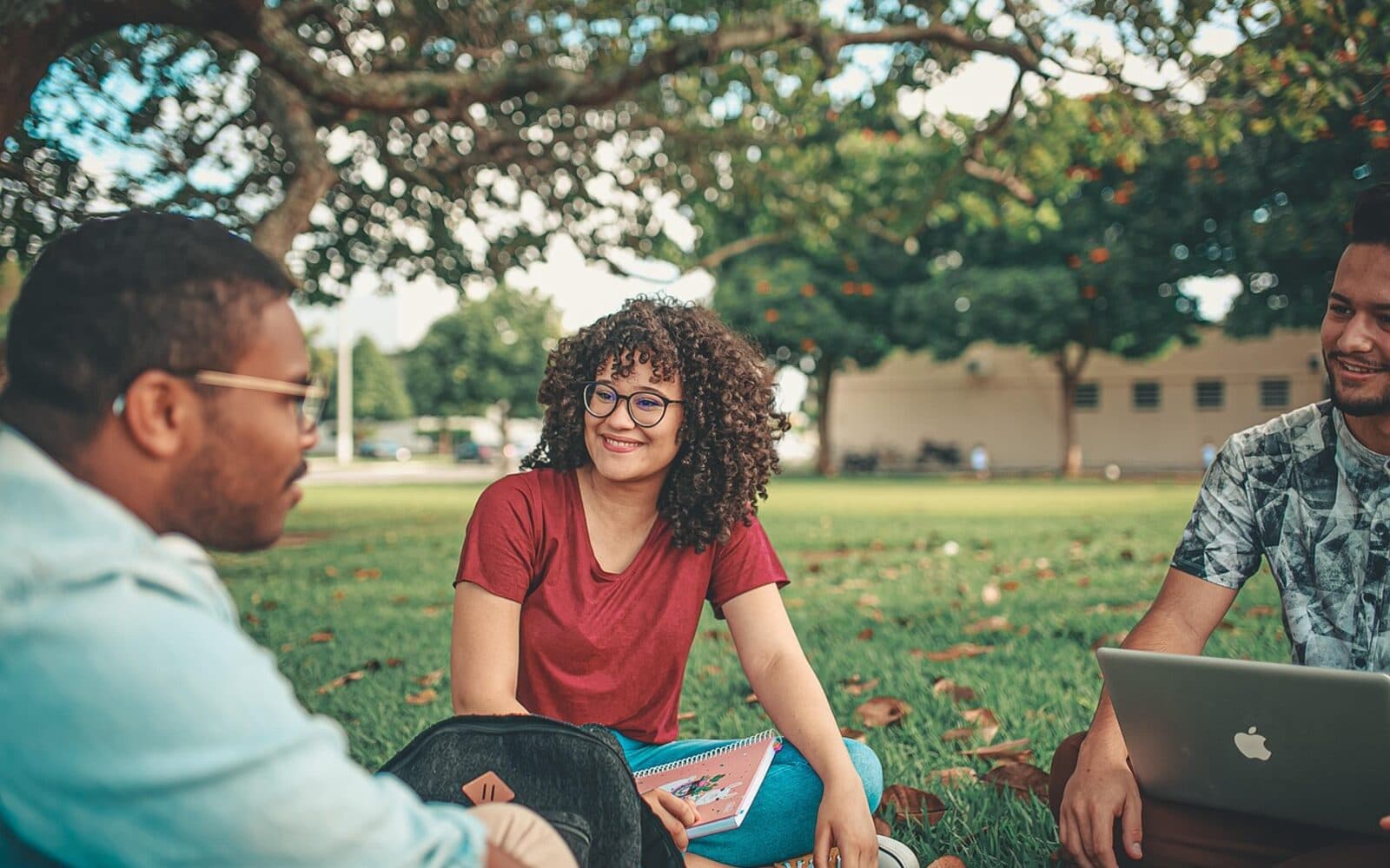 Concept image of three university students chatting and laughing together on the lawn of a sunny campus, with a laptop and notebook nearby.